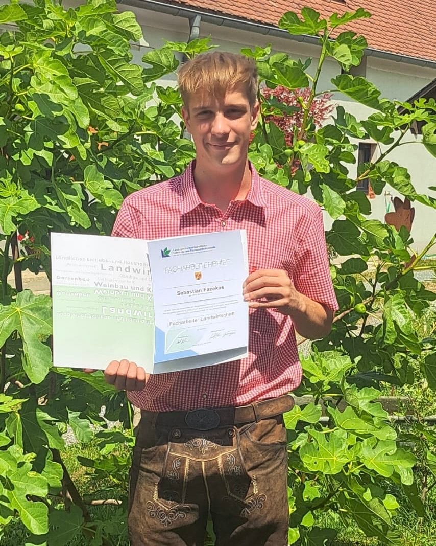 A young man in a red checkered shirt poses with a certificate. The certificate reads Facharbeiterbrief Landwirtschaft. He stands in front of a green leafy plant.