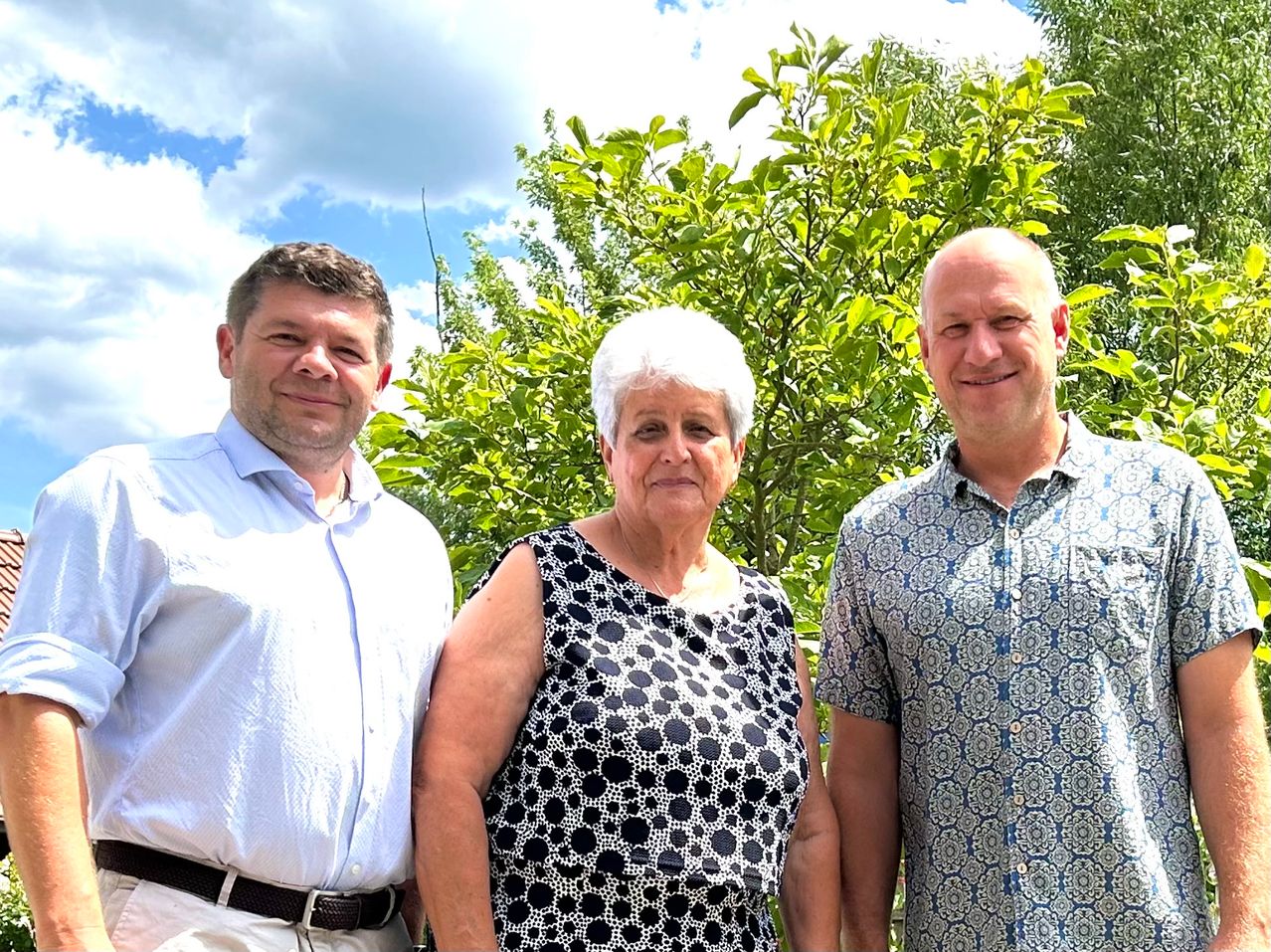 Three people are standing close to each other, smiling, and posing for a photo in front of a tree.