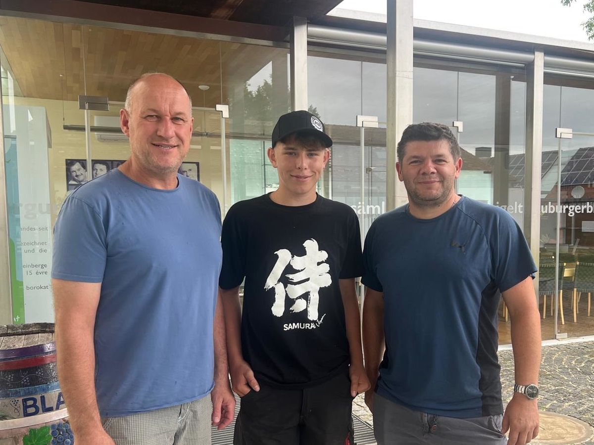 Three men pose for a photo outside a building with glass walls, smiling and standing close together. One wears a black hat and shirt with 'Samurai' written on it.