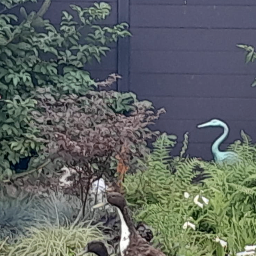 A garden scene with lush greenery, a tree, and a bird statue on the right. The garden is surrounded by a wooden fence.