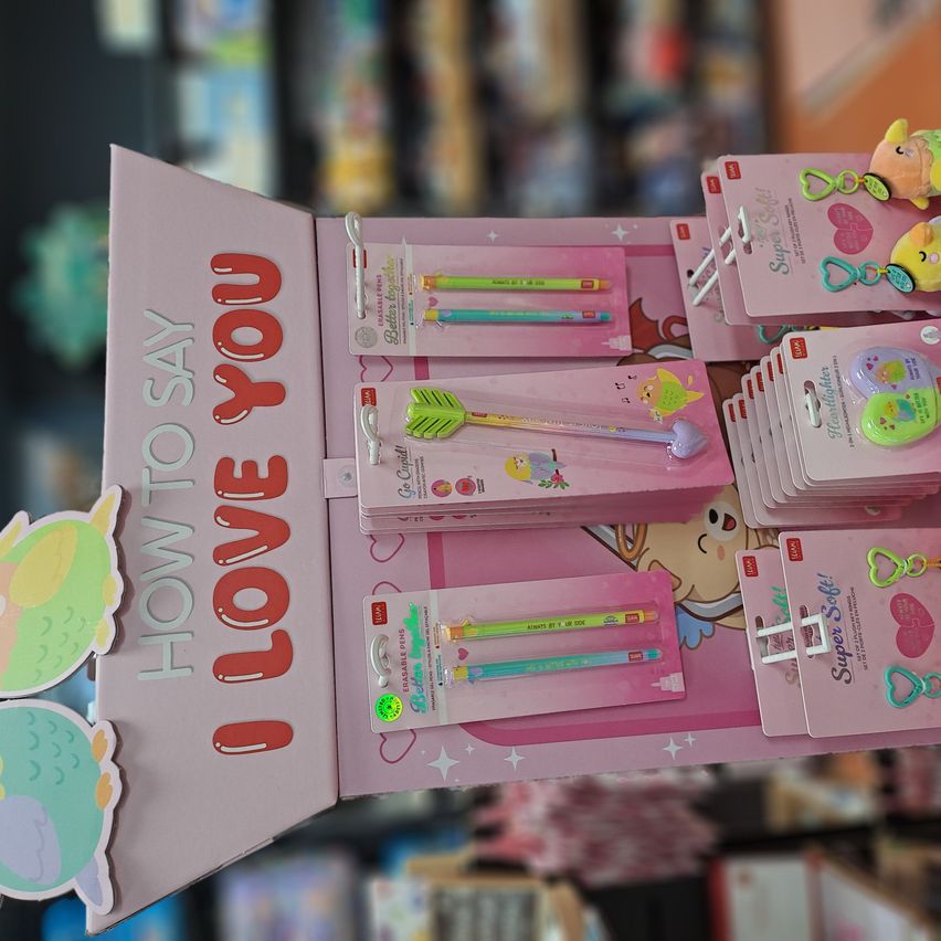 Eine offene rosafarbene Box zeigt verschiedene Schreibwaren, darunter Bleistifte, Stifte und Schlüsselanhänger. Die Box trägt den Text 'Wie man sagt, ich liebe dich' und enthält farbenfrohe und thematische Produkte.