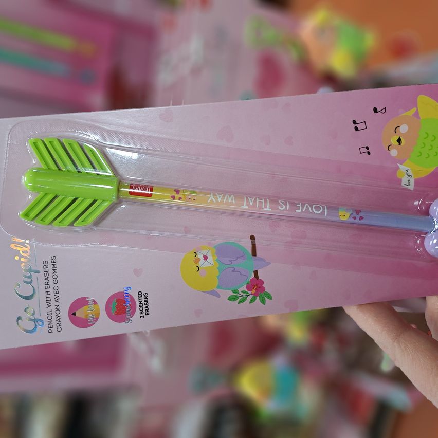 Eine rosa Verpackung zeigt einen grünen Pfeilstift mit einem Stift und einem Radiergummi, in Plastik verpackt. Die Verpackung zeigt einen fröhlichen Charakter mit Musiknoten und einem Blumendesign.