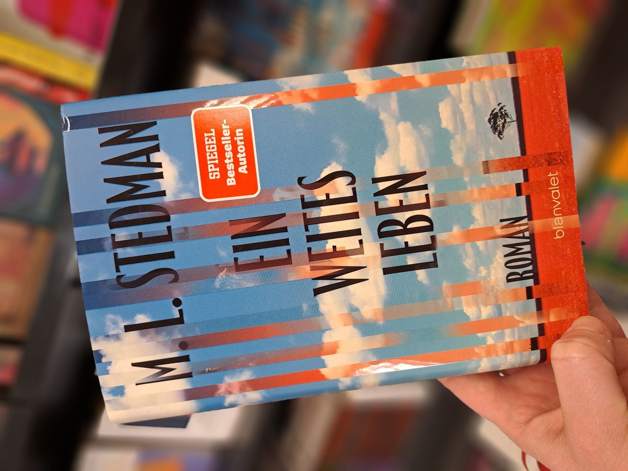A person holds a book titled 'Ein Weites Leben' by M. L. Stedman. The cover is blue and orange, with clouds in the background.