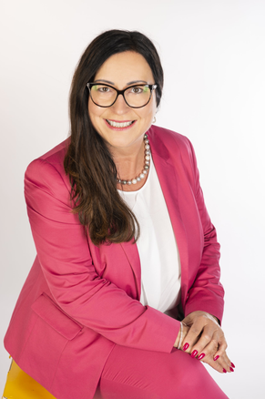 Eine Frau mit langen dunklen Haaren, Brille und einem rosa Blazer über einem weißen Hemd, lächelt und posiert für ein Foto.
