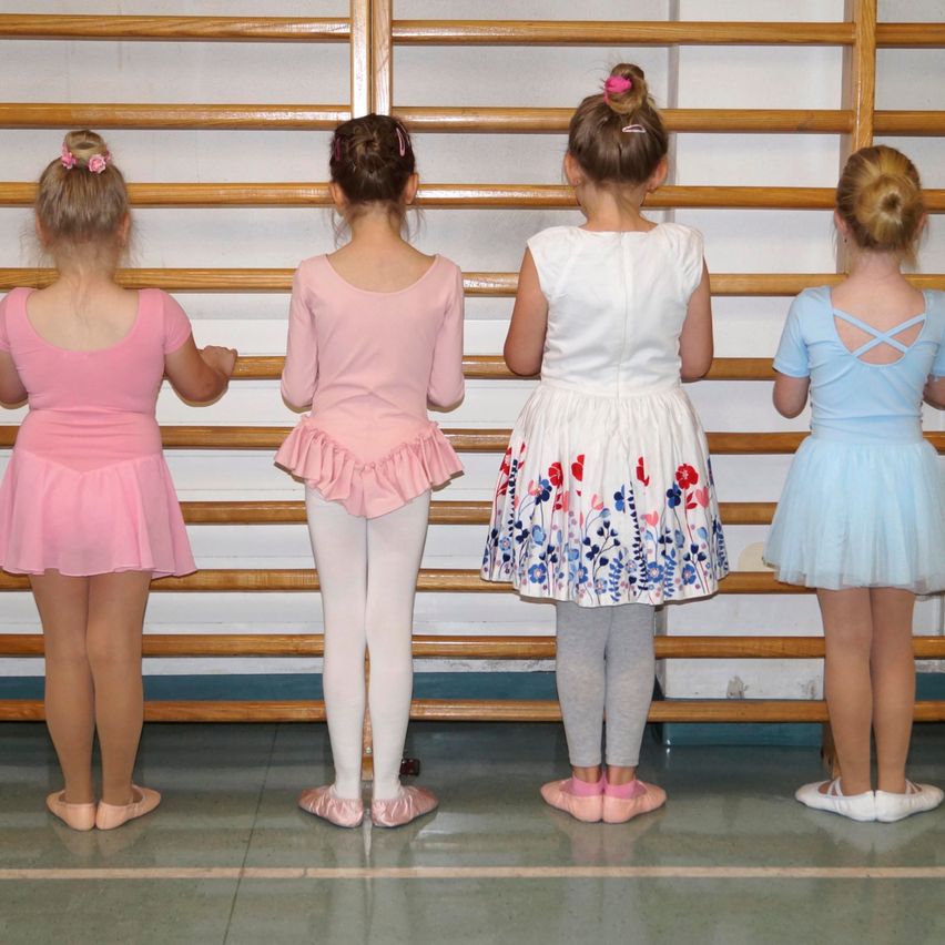 Four young girls in ballet attire stand in a row in a dance studio, facing away from the camera. Each wears different colored dresses and ballet shoes.