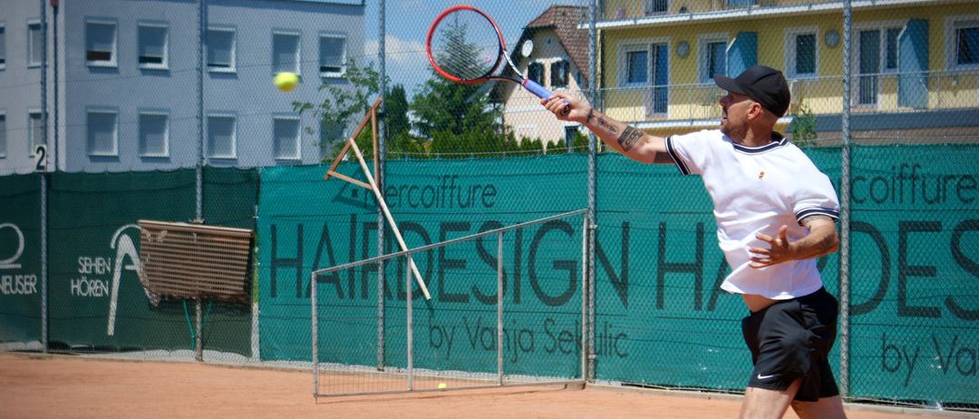 Ein Mann spielt Tennis auf einem Sandplatz und schlägt einen Ball mit einem Schläger. Der Platz ist von einer grünen Banner und einer Metallbarriere umgeben.