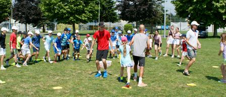 Eine Gruppe von Kindern in Mützen und Turnschuhen rennt über ein Grasfeld. Ein Mann in einem roten T-Shirt und blauen Schuhen geht in der Nähe. Ein Mädchen in einer rosa Mütze und ein Mädchen in einem blauen Kleid stehen in seiner Nähe.