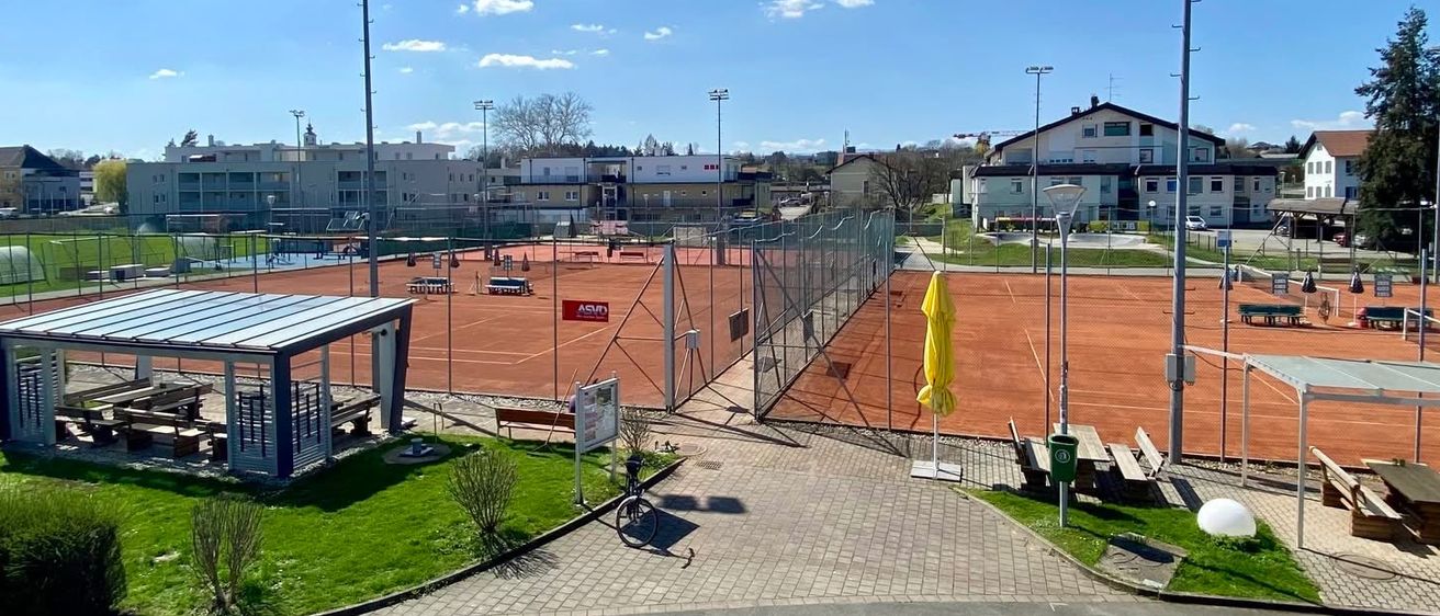 Ein Tennisplatz-Komplex mit mehreren Plätzen, einer Laube und Bänken. Bäume und Straßenlampen sind vorhanden. Der Himmel ist blau mit Wolken.