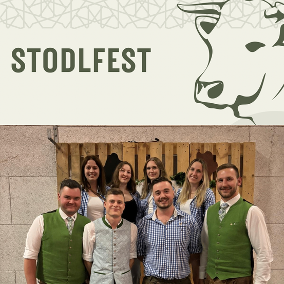 Eine Gruppe von Menschen in traditioneller bayerischer Kleidung, einschließlich Männer und Frauen, stehen vor einer Holzwand und lächeln für ein Foto.