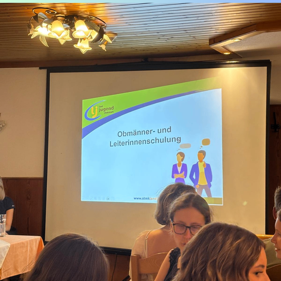 Eine Gruppe von Menschen sitzt in einem Raum und beobachtet eine Präsentation auf einer Projektionsleinwand. Die gezeigte Folie hat eine Grafik von zwei Personen mit dem Text 'Oebuenner-und Leiterinnenschulung'.