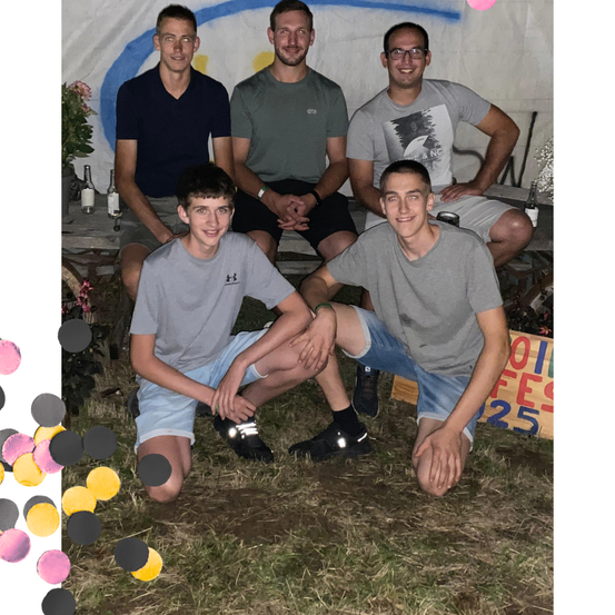 Fünf junge Männer posieren für ein Foto im Freien. Sie sitzen auf dem Gras, mit einem bemalten Hintergrund vor ihnen.