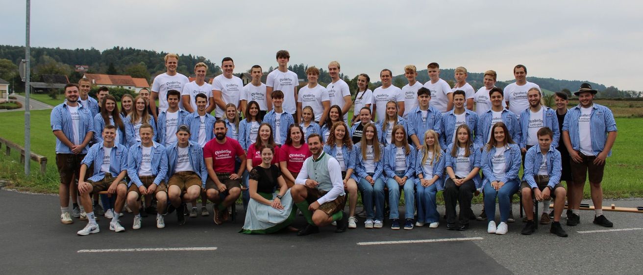 Eine Gruppe von Menschen in blauen und weißen Hemden und Kleidern posiert für ein Gruppenfoto. Einige von ihnen sitzen auf dem Boden. Sie stehen vor einem weißen Gebäude mit einem Berg in der Ferne.