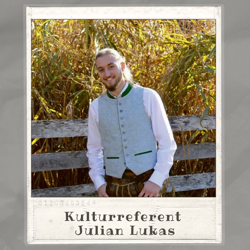 Ein lächelnder Mann steht vor einem Maisfeld, trägt einen Kilt, ein weißes Hemd und eine graue Weste. Sein Name, Julian Lukas, wird unten angezeigt.
