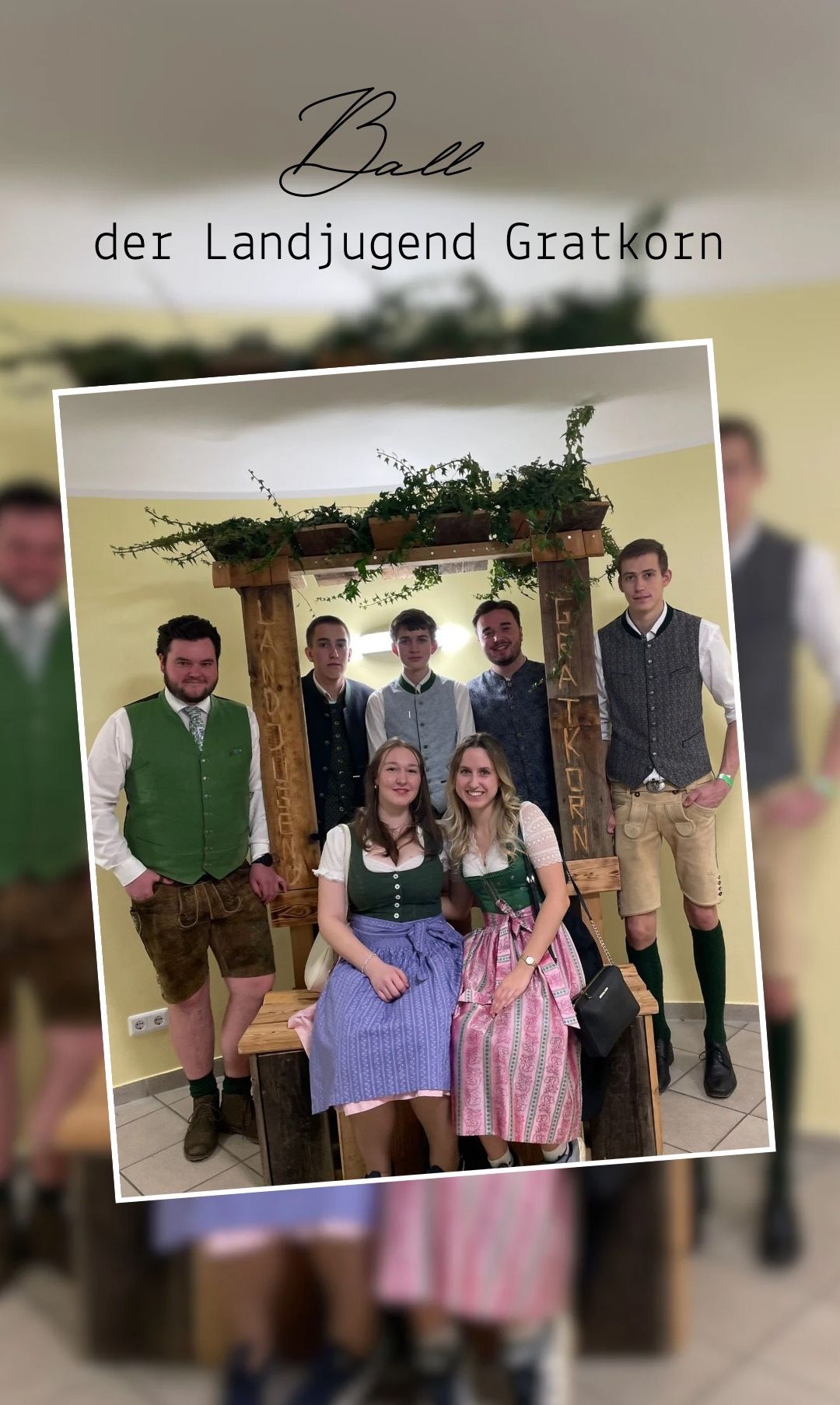 Gruppenfoto von Menschen in traditioneller bayerischer Kleidung, die vor einer Holzstruktur mit Pflanzen und dem Wort 'Korn' darauf stehen. Zwei Frauen sitzen auf einer Bank, und zwei Männer stehen hinter ihnen.