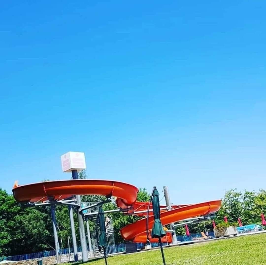 Eine orangefarbene Wasserrutsche unter einem klaren blauen Himmel mit einem grünen Regenschirm und Bäumen im Hintergrund.