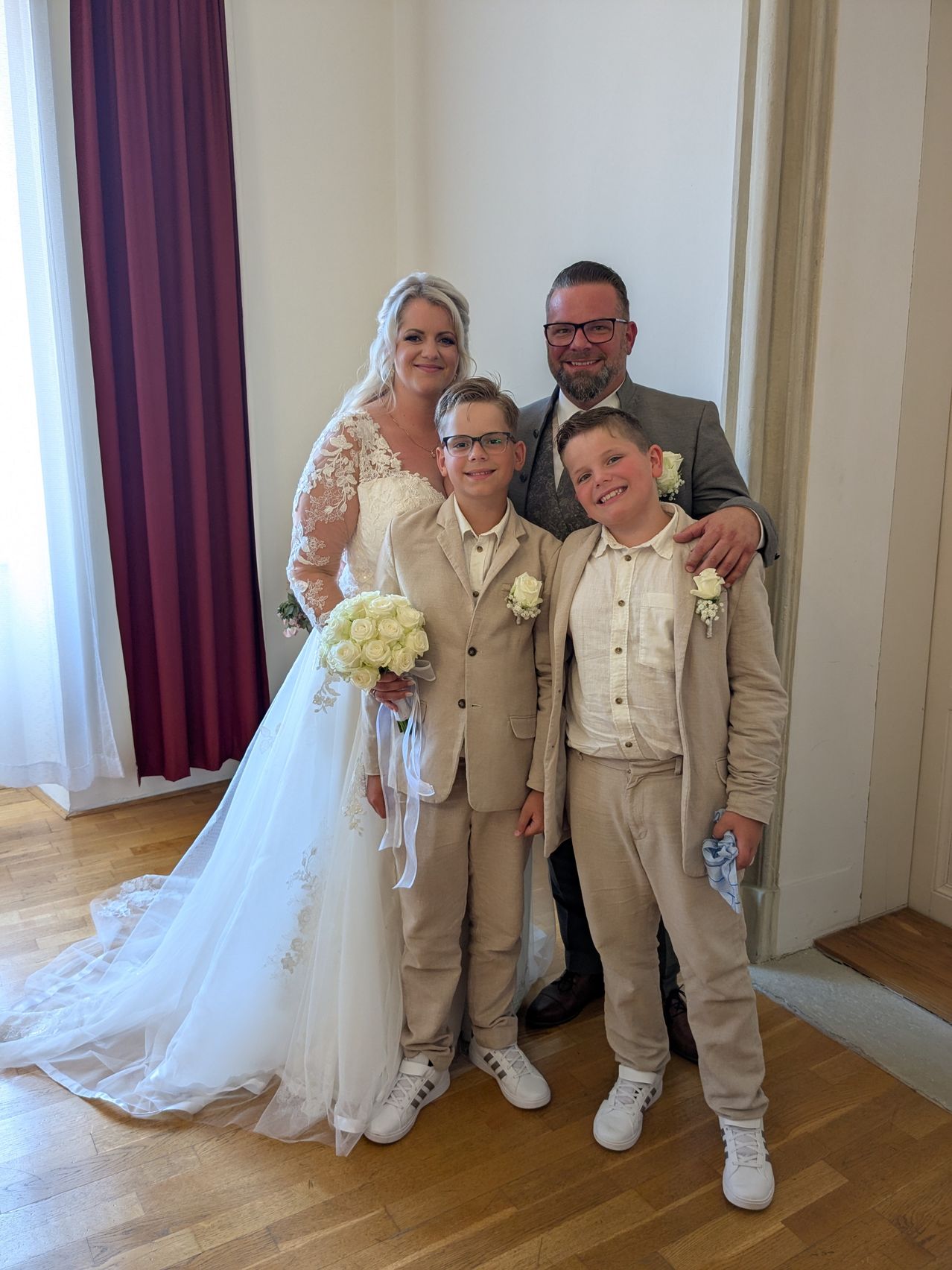 Eine frisch verheiratete Frau in einem weißen Kleid hält einen Blumenstrauß und steht neben zwei lächelnden Jungen in Anzügen und einem Mann mit Brille, die alle für ein Foto in einem Raum posieren.