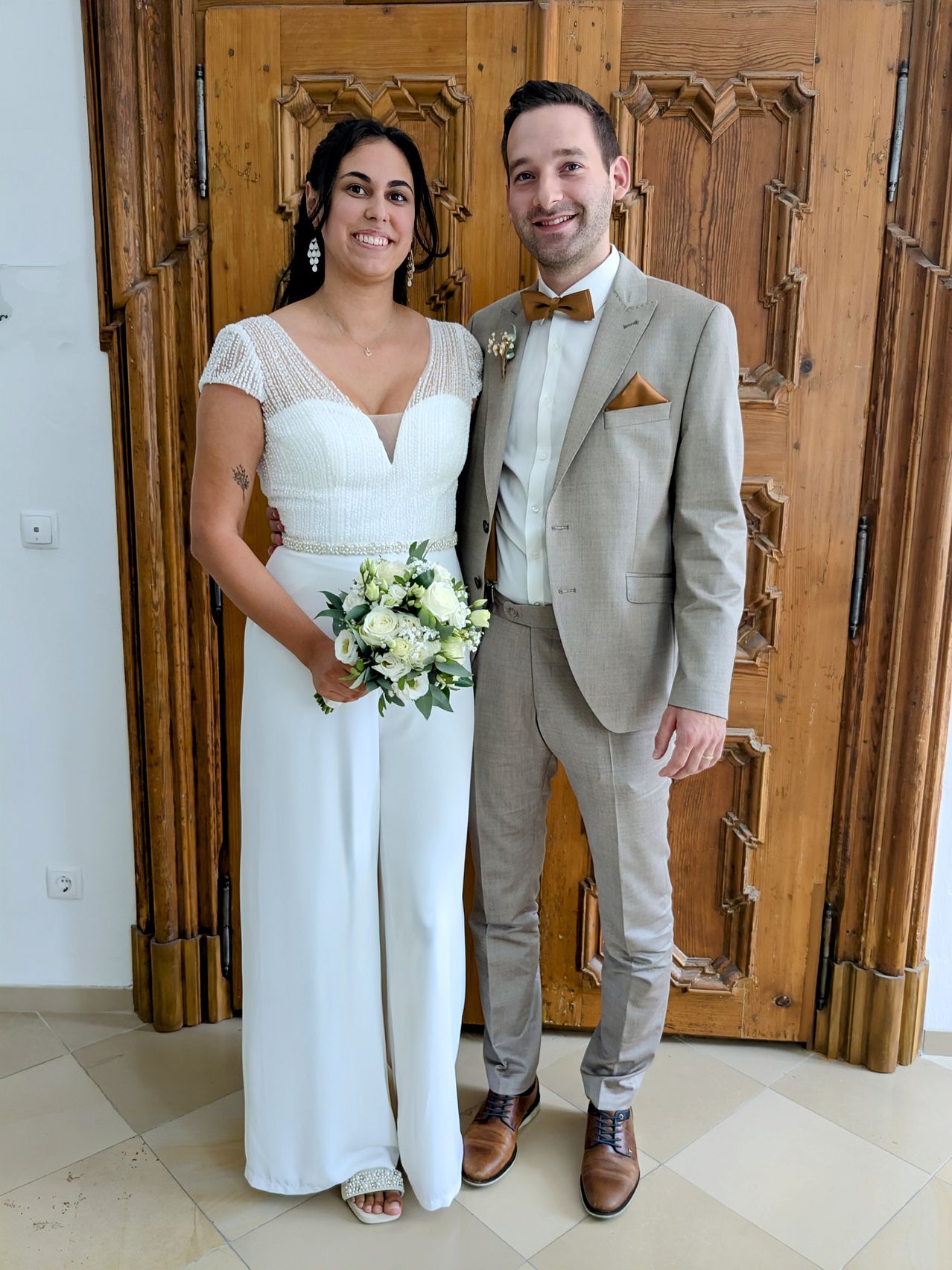 Ein frisch verheiratetes Paar steht lächelnd vor einer verzierten Holztür. Die Braut trägt ein weißes Kleid und hält eine Blumenstrauß. Der Bräutigam ist in einem hellen Anzug mit einer Fliege gekleidet.