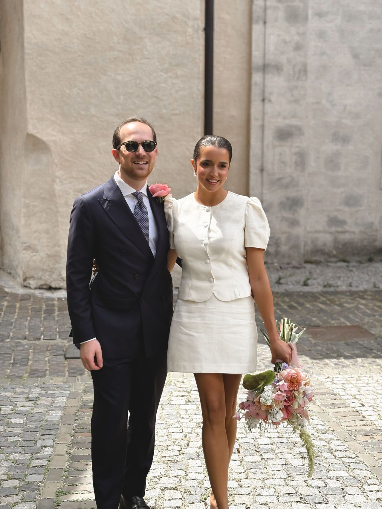 Ein lächelndes Paar steht auf einem Kopfsteinpflasterweg, der Mann in einem Anzug mit einer rosa Blume, die Frau in einem weißen Kleid mit einem Blumenstrauß.
