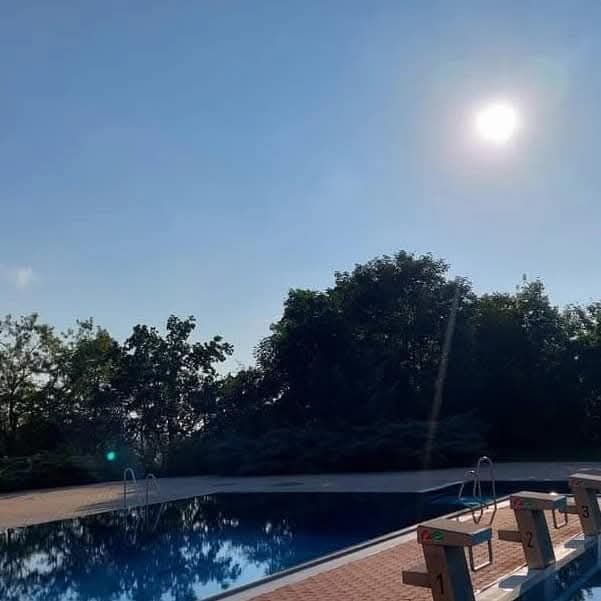 Ein Freibad unter einem klaren blauen Himmel, umgeben von Bäumen. Die Sonne scheint hell. Der Pool ist ruhig mit Bahnmarkierungen, die mit 1, 2 und 3 nummeriert sind.