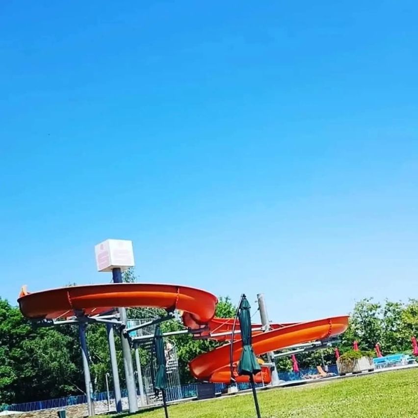 Ein Wasserpark im Freien mit blauem Himmel im Hintergrund. Eine große orangefarbene Wasserrutsche mit einem geschlossenen Schirm in der Nähe. Bäume und ein Zaun umgeben das Gebiet.