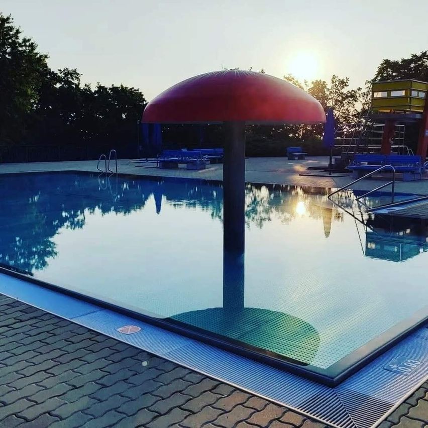 Ein roter, pilzförmiger Gegenstand steht in der Mitte eines Schwimmbeckens und reflektiert den Himmel und die Bäume. Bänke und geschlossene Schirme befinden sich um das Becken herum.