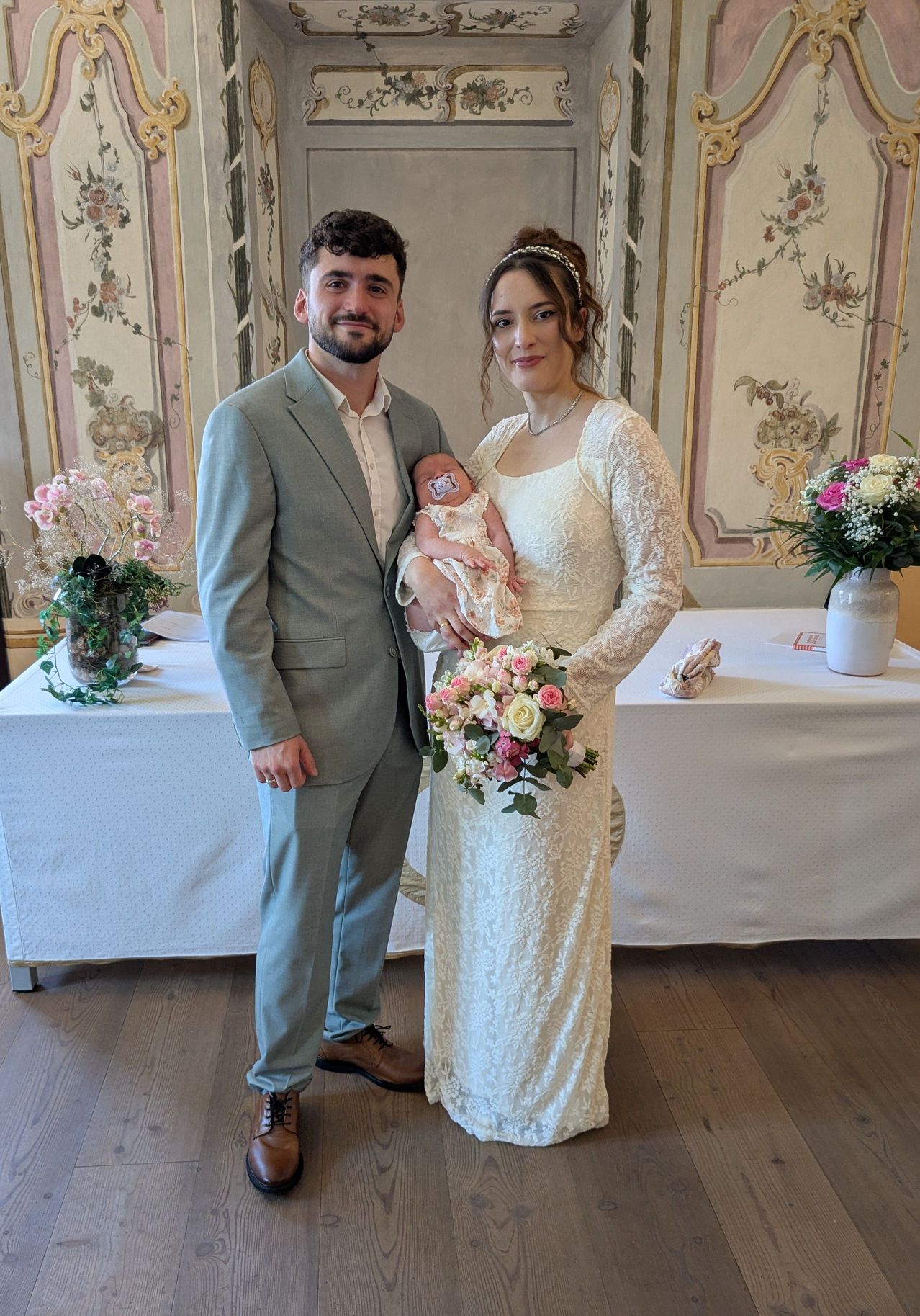 Ein Paar steht in einem Raum mit Blumendekor. Die Frau hält ein Baby und trägt ein Hochzeitskleid. Der Mann trägt einen grauen Anzug. Hinter ihnen steht ein Tisch mit Blumen und einer Vase.