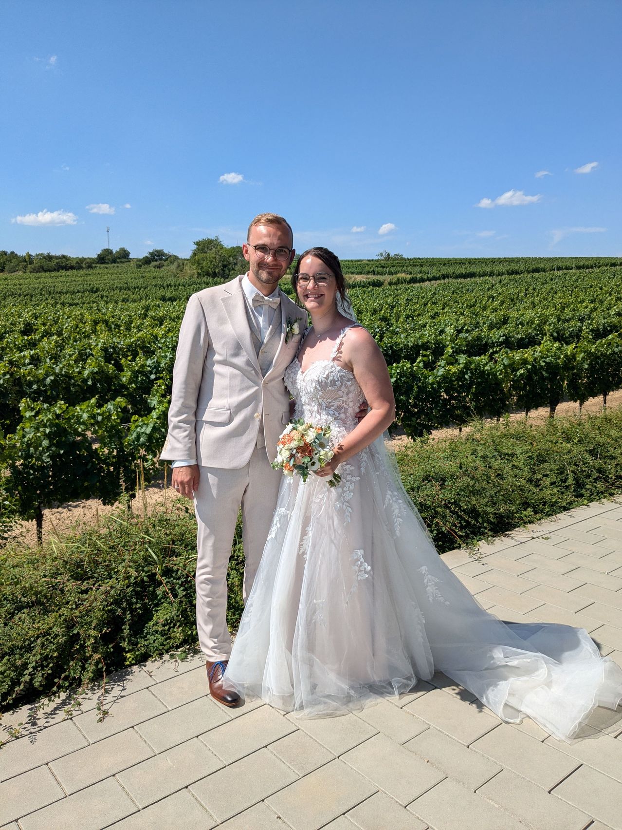 Ein frisch verheiratetes Paar steht in einem Weinberg mit klarem blauem Himmel. Der Bräutigam trägt einen hellbraunen Anzug und die Braut ein Hochzeitskleid mit langem Schleier.
