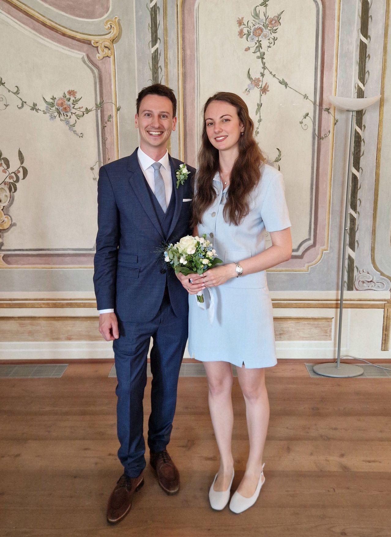 Ein Mann und eine Frau stehen eng beieinander, lächeln für ein Foto, wobei die Frau einen Blumenstrauß hält. Sie sind in hellblauen Anzügen gekleidet und stehen auf einem Holzboden vor einer Wand mit Blumengemälden.