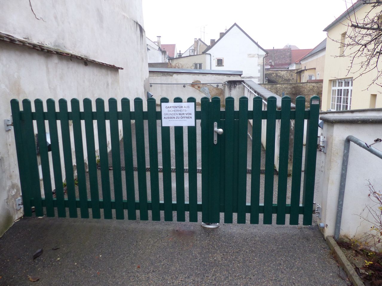 Ein grünes Tor mit einem Schild, das 'GARTENTOR AUS SICHERHEITS-GRUNDEN NUR VON AUSSEN ZU OFFNEN' steht, ist geschlossen. Das Tor befindet sich auf einer gepflasterten Straße mit Gebäuden dahinter.