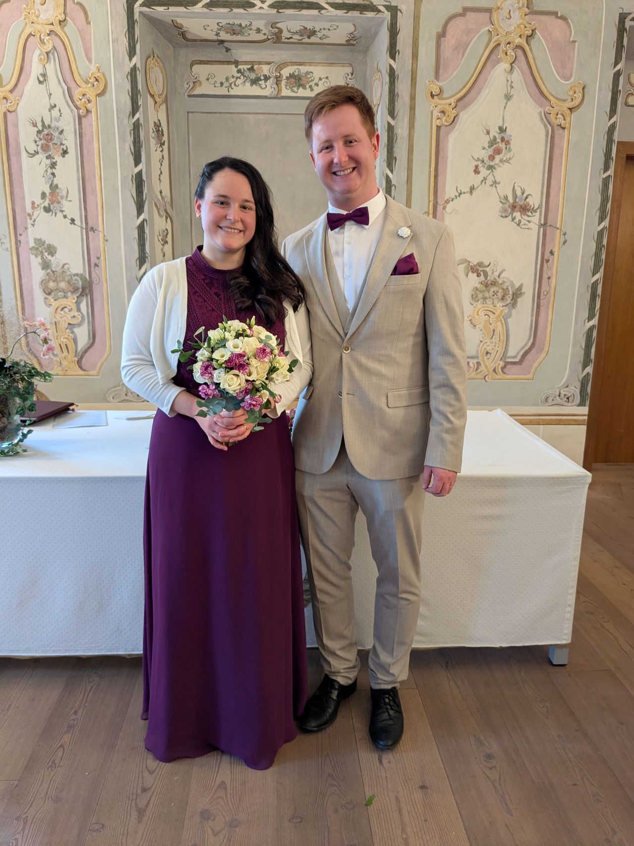 Ein Mann und eine Frau stehen nah beieinander in einem Raum und lächeln für ein Foto. Die Frau hält einen Blumenstrauß. Hinter ihnen steht ein Tisch mit einer weißen Tischdecke.