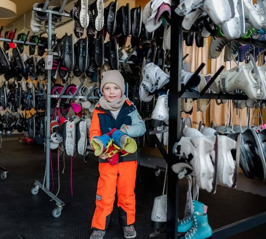 Ein kleiner Junge steht vor einem Schuhregal und hält ein Paar Schlittschuhe. Er trägt eine Mütze und einen orangefarbenen und blauen Schneeanzug.