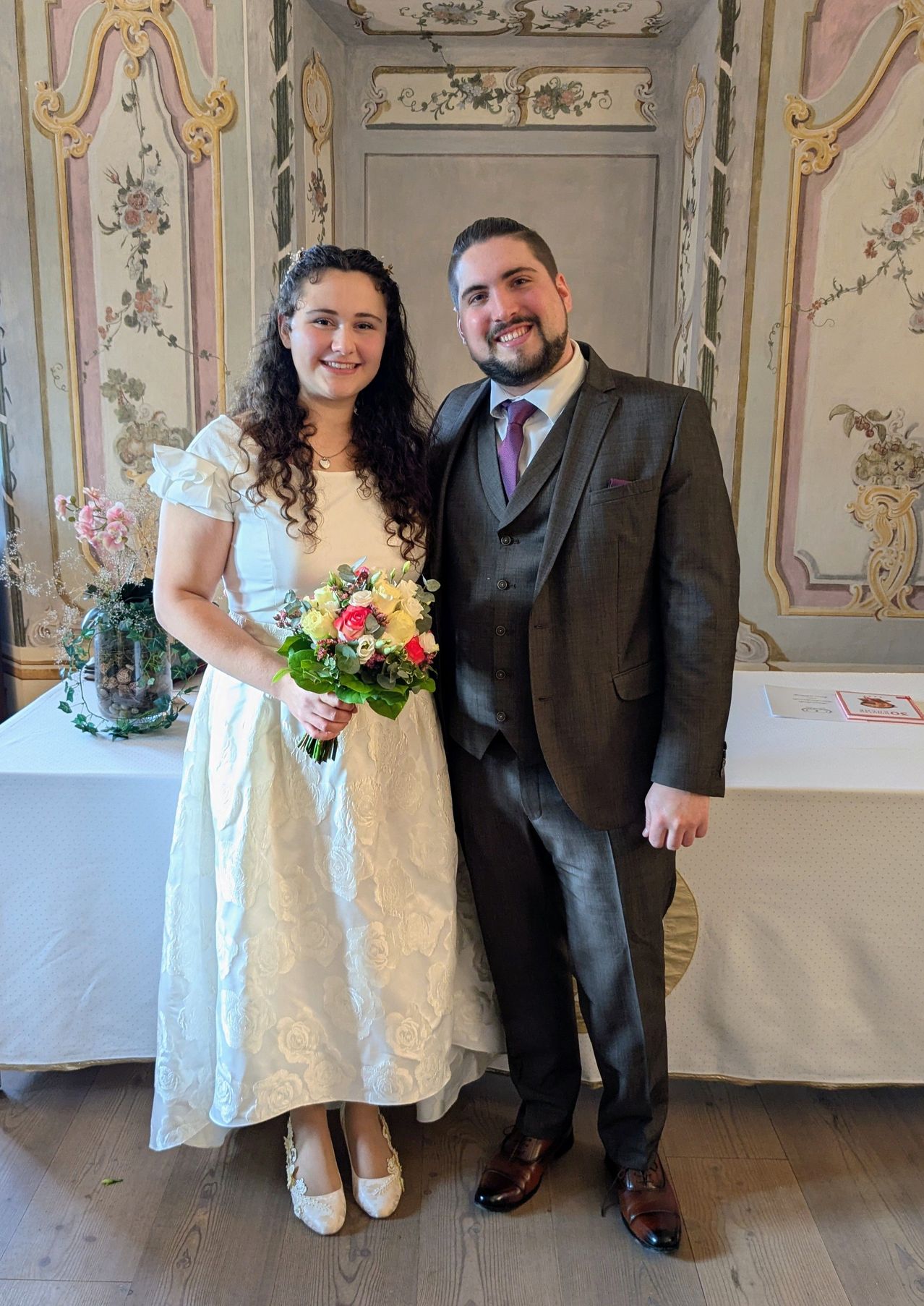 Ein frisch verheiratetes Paar steht lächelnd vor einer kunstvoll verzierten Wand mit Blumendekorationen für ein Foto. Die Frau hält einen Blumenstrauß in der Hand.