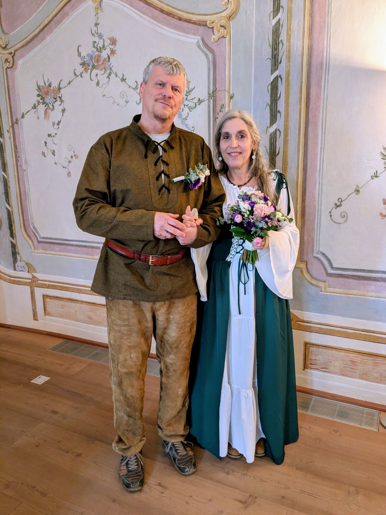 Ein Mann und eine Frau in mittelalterlicher Kleidung stehen lächelnd in einem Raum mit Blumenwanddesigns.