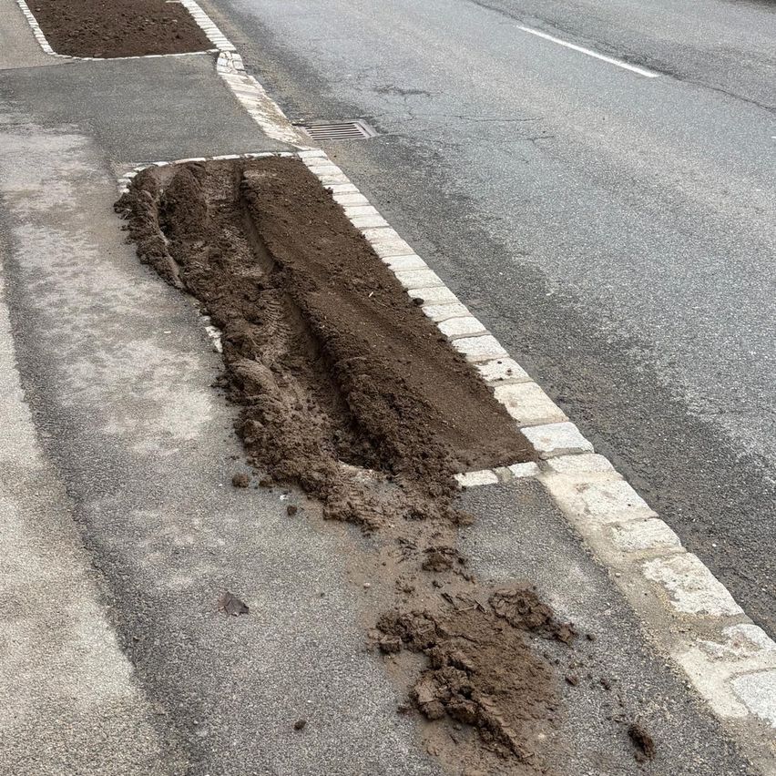 Eine Straße hat einen frisch gegrabenen Graben entlang ihres Bordsteins, der Erde freilegt. Schmutz ist auf der Straßenoberfläche verstreut.