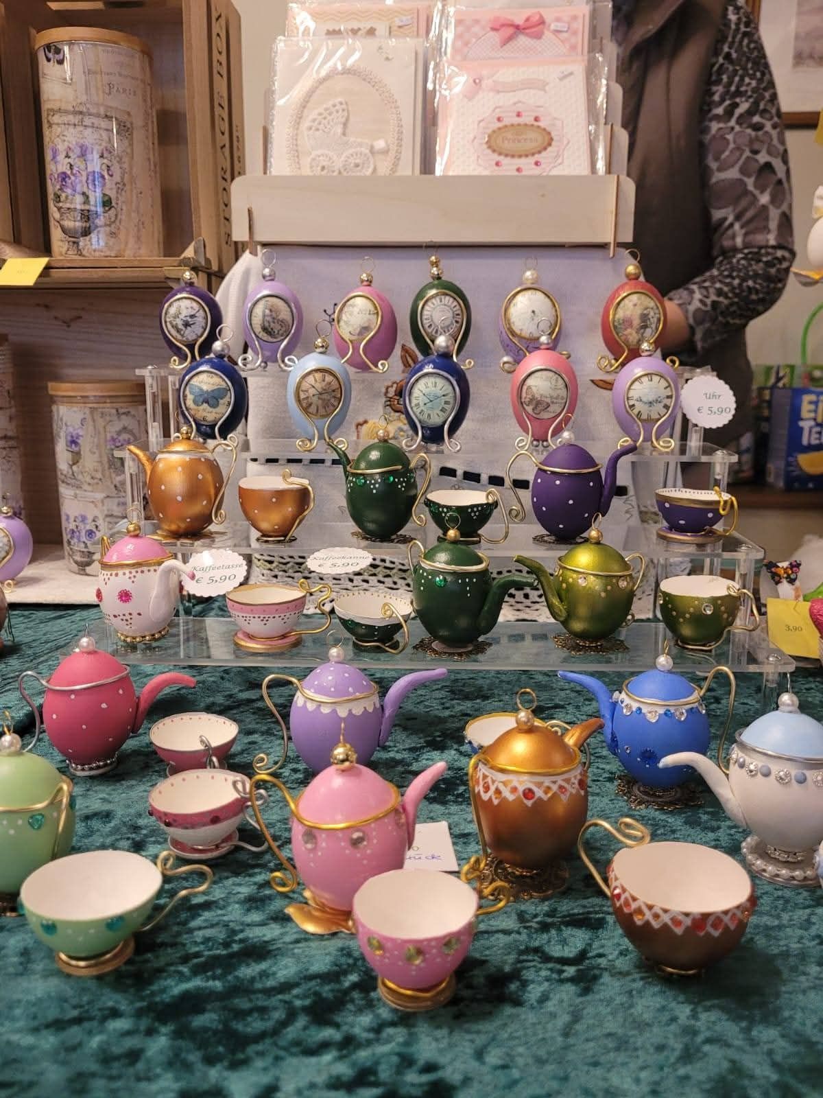 Eine Ausstellung von dekorativen Teekannen und Tassen auf einem Tisch mit einem türkisen Tuch. Jede Teekanne hat ein einzigartiges Design und eine Farbe, wobei einige Uhren auf ihren Deckeln haben. Es gibt auch kleinere Tassen und Untertassen. Im Hintergrund steht eine Person in der Nähe von Regalen mit weiteren Gegenständen.