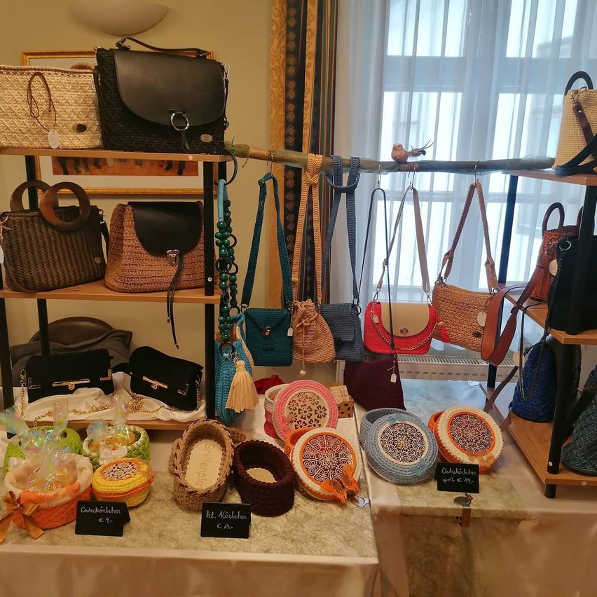 Eine Auswahl an handgefertigten Taschen und Körben wird auf Regalen und einem Tisch ausgestellt, darunter geflochtene und gestrickte Artikel. Jeder Artikel hat ein Preisschild und einige sind in Plastik verpackt.