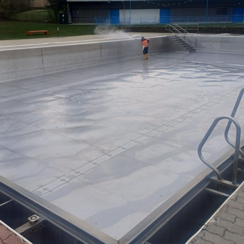 Eine Person reinigt einen leeren Pool mit einem Schlauch, trägt eine Warnweste und Stiefel, vor einem Gebäude mit blauen Türen und Geländern.