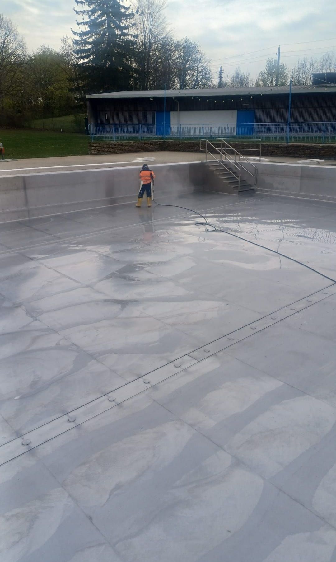 Ein Arbeiter in einer orangefarbenen Jacke und gelben Stiefeln steht in einem Pool und hält einen Schlauch, der Wasser auf die Oberfläche des Pools sprüht. Der Pool ist von einer Betonbarriere umgeben und hat eine Treppe, die zum Wasser führt.