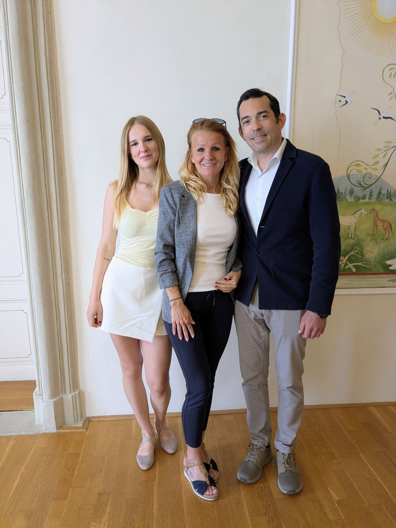 Three people stand close together in a room with wooden floors and white walls. A woman on the right wears a suit. The woman in the middle has a gray jacket.
