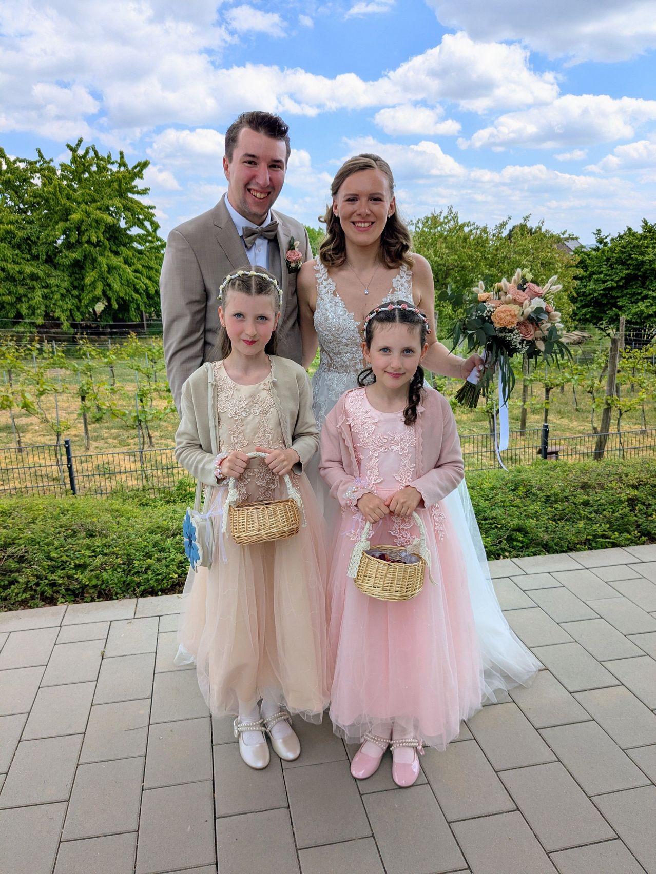 A wedding party, a man, a woman, and two young girls stand on a tiled pathway in a garden. They are all smiling.