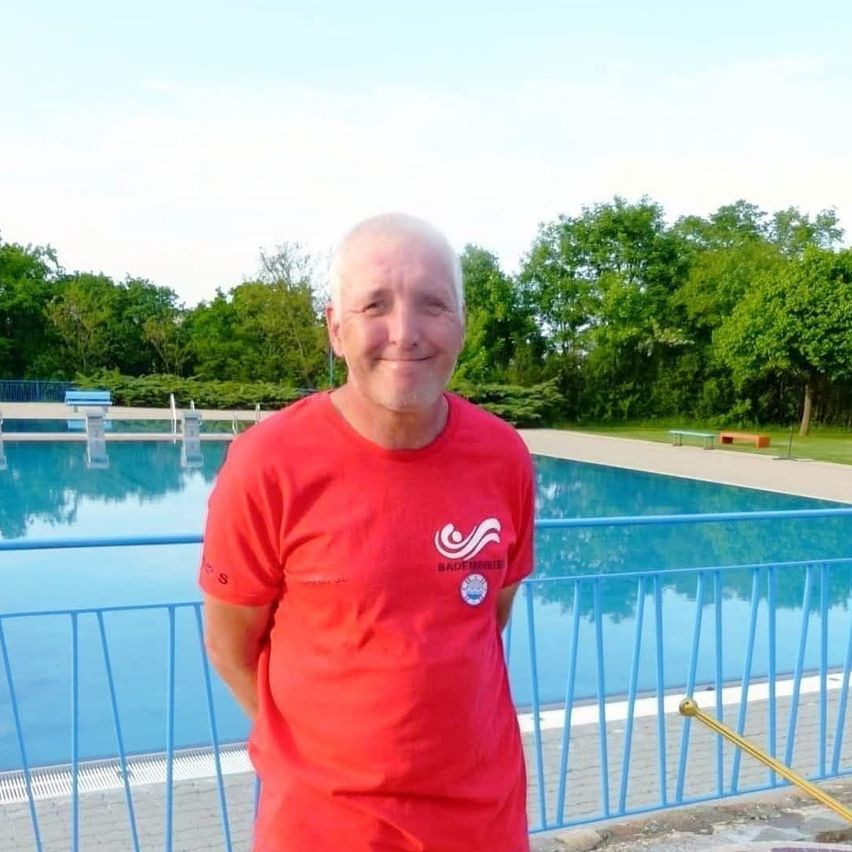 A smiling man in a red shirt stands near a blue fence around an outdoor swimming pool with trees in the background.