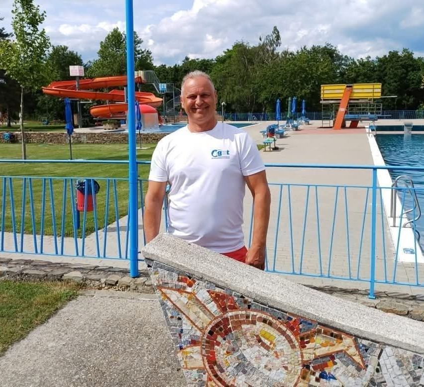 A man stands at a blue fence in a park with a pool, smiling. Behind him, there is an orange water slide and a large yellow slide.