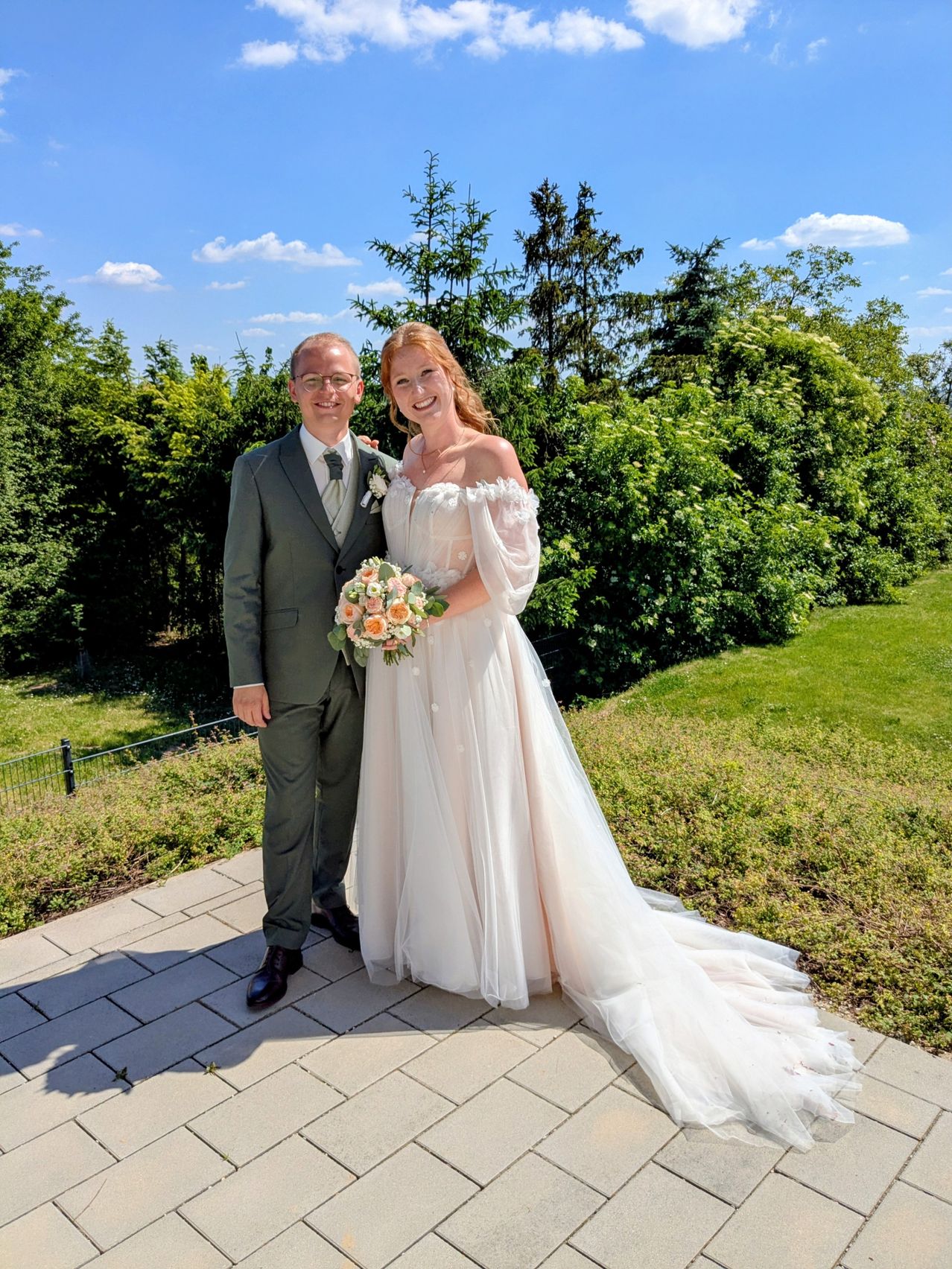 Ein Mann und eine Frau in Hochzeitskleidung stehen lächelnd zusammen auf einem gepflasterten Gehweg. Die Frau hält einen Blumenstrauß, und sie sind von Grünpflanzen und einem Zaun umgeben.