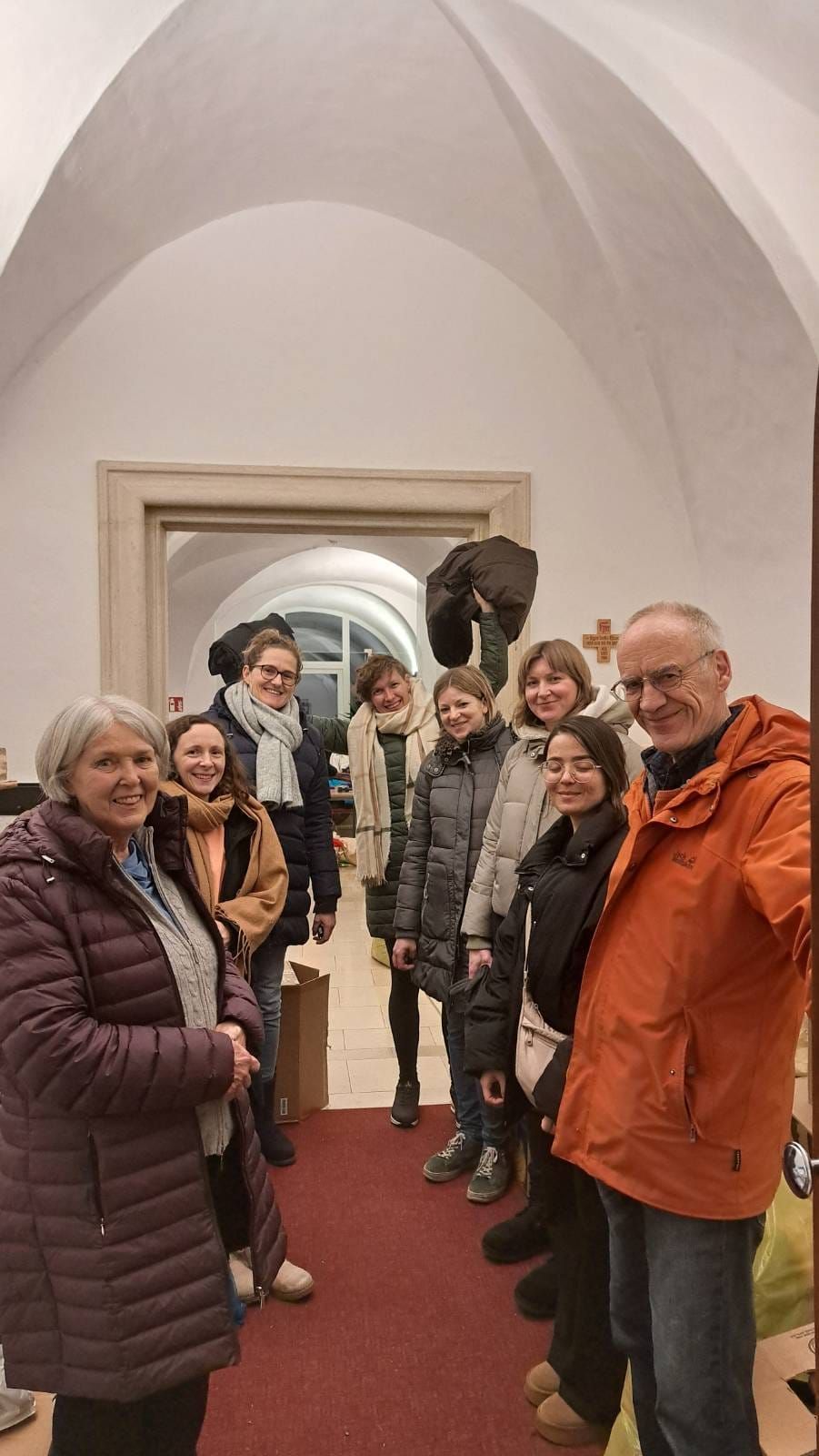 A group of smiling people in warm clothing stand in a hallway with a cross on the wall, posing for a photo.