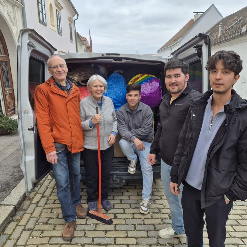 Five people stand near a van on a cobblestone street, smiling. The van's back door is open, and they are holding bags and a broom. Behind them are buildings with traditional architecture.