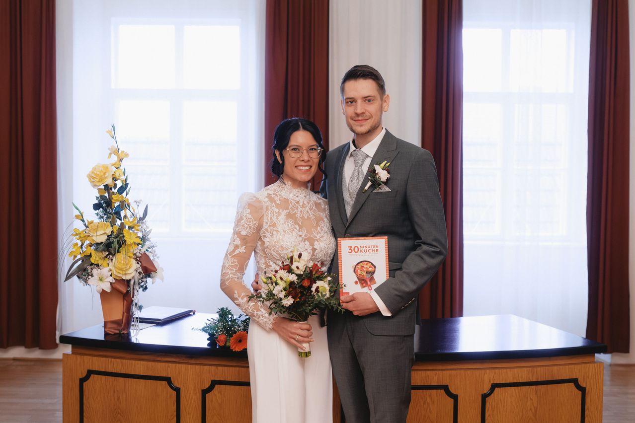 Ein Mann und eine Frau stehen nah beieinander in Hochzeitskleidung und halten ein Buch mit dem Titel '30 Minuten Kuche'. Dahinter befinden sich Blumen und ein hölzerner Schreibtisch.