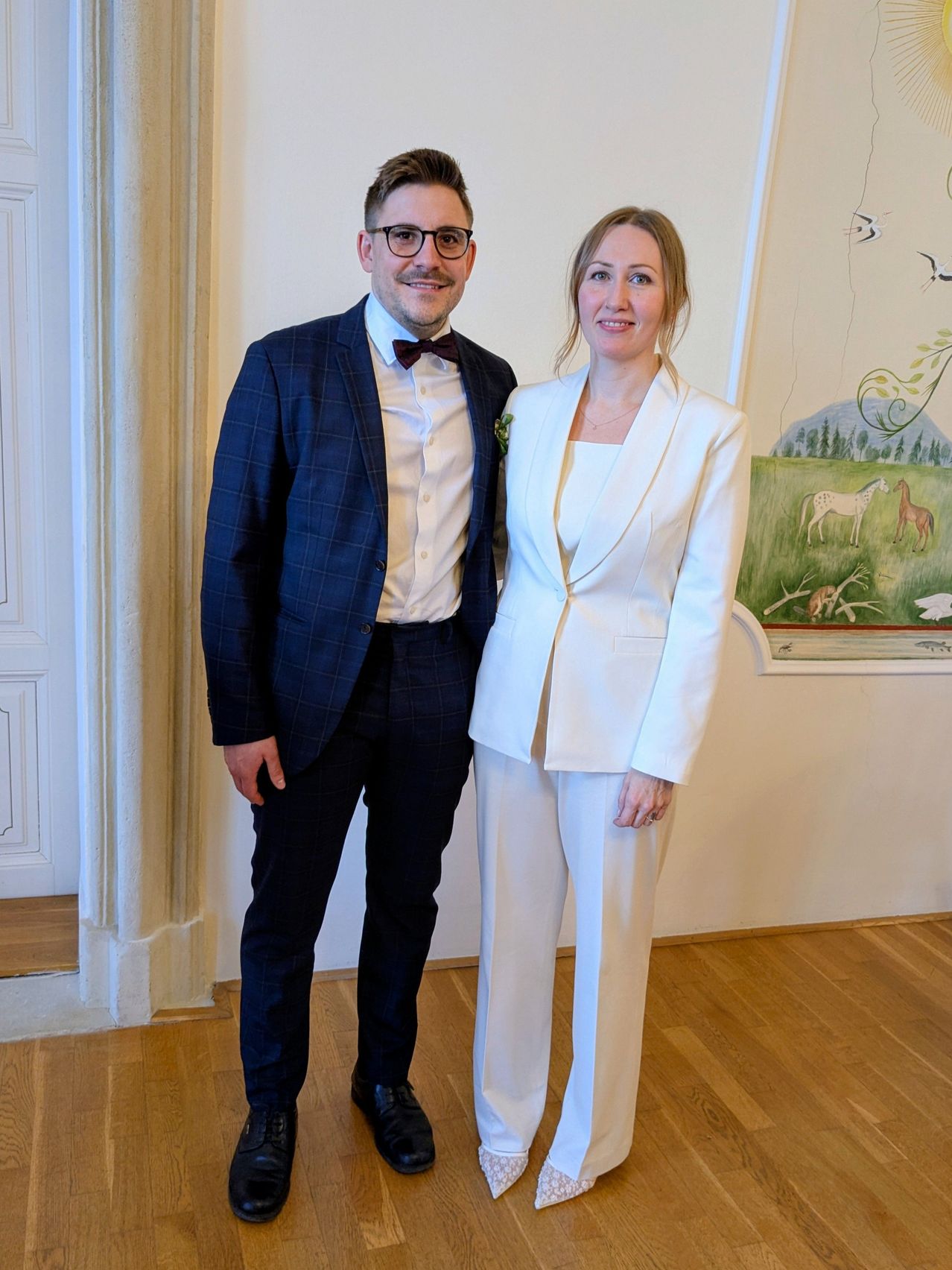 Ein Mann und eine Frau stehen eng beieinander. Sie lächeln. Der Mann trägt einen Anzug mit einer Fliege. Die Frau trägt einen weißen Anzug. Sie stehen vor einer Wand mit einem Gemälde.
