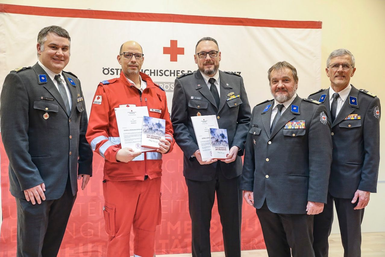 Fünf Männer in Uniformen stehen nebeneinander. Der Mann in der Mitte hält ein Zertifikat und ein Buch. Die Männer links und rechts halten nur Zertifikate. Ein Banner mit einem roten Kreuz ist hinter ihnen.