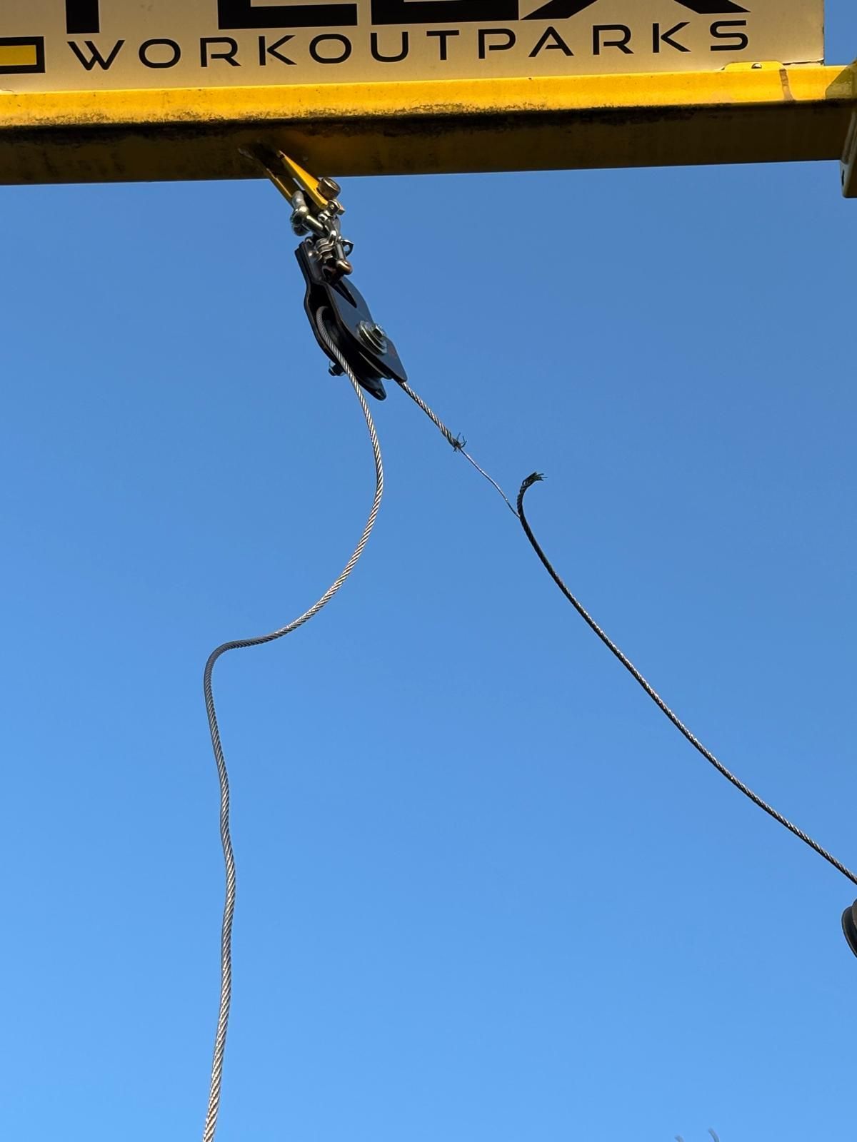 Ein Kran mit gelbem Haken und Flaschenzugsystem hängt in der Luft mit einem Seil, vor einem klaren blauen Himmel.