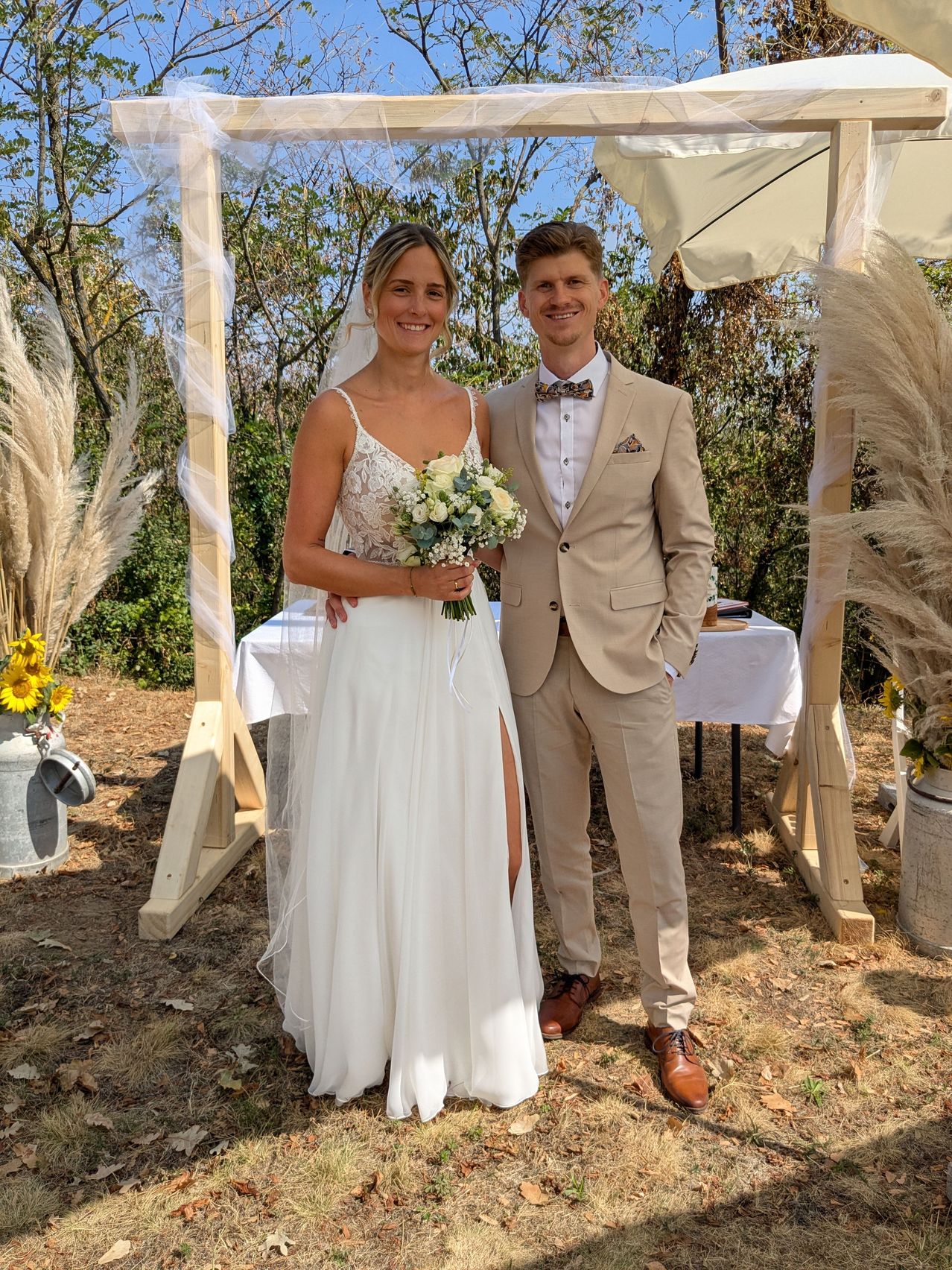 Ein frisch verheiratetes Paar steht unter einem rustikalen Holzbogen, die Braut in einem weißen Kleid und der Bräutigam in einem hellbraunen Anzug, beide lächeln in die Kamera.